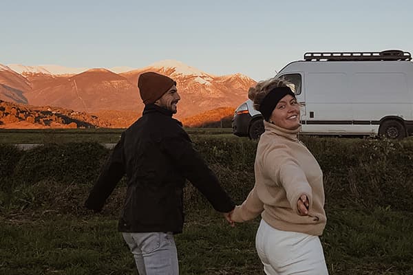 My new owners: Iris & Max - irisandmax 06/2022 A young couple, @irisandmax, holding hands and smiling in front of their converted Renault Master van. The scenic backdrop features snow-capped mountains and a golden sunset, embodying the essence of vanlife and outdoor exploration.
