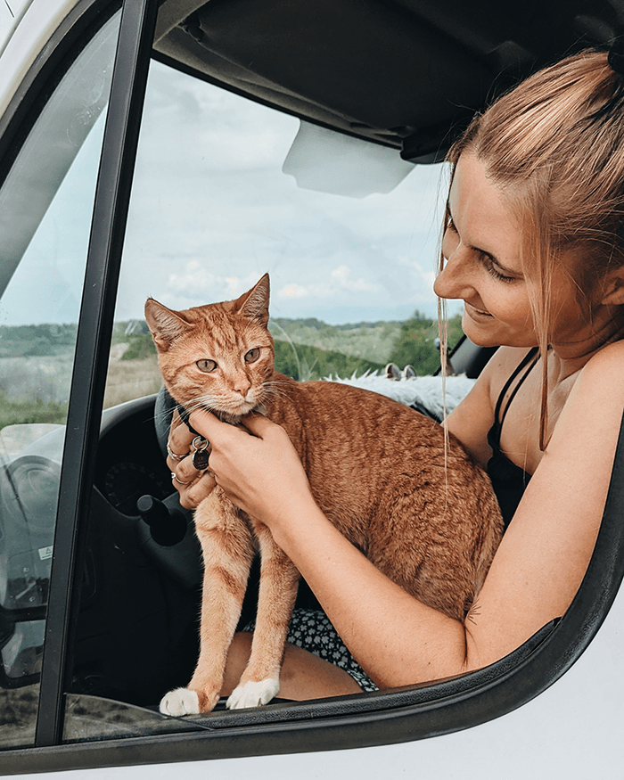 More relaxed & happier than ever - @chanti.and.ollie 12/2022 Chantal from @chanti.and.ollie smiles while holding her ginger cat Ollie on her lap in the driver’s seat of their white Opel Movano campervan, parked in nature during a European vanlife journey.
