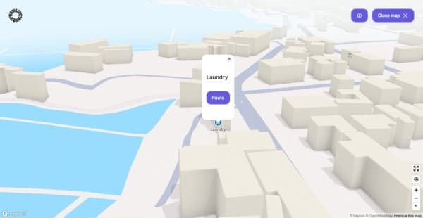 A laundromat near you - vanlifezone.com/map A 3D interactive city map from Vanlifezone, featuring a simplified representation of an urban layout with block-like buildings and blue water. A pop-up on the map highlights a 'Laundry' location with a 'Route' button to guide users to the service. The interface includes a 'Close map' button and map navigation tools to the right, as well as a Mapbox and OpenStreetMap attribution in the lower right corner, indicating the map's source.