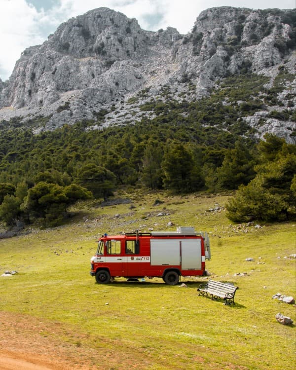 Hummel parked in nature - @hummel_die_feuerwehr 06/2022 A red firetruck, @hummel_die_feuerwehr, converted into a camper parked in a grassy field at the base of a rocky mountain, showcasing the vanlife experience in a remote and rugged natural setting on Crete’s south coast.