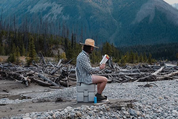 When nature calls - @christiandelatorre_ 09/2022 Christian de la Torre (@christiandelatorre_) sits on a portable toilet in a tranquil wilderness setting, reading a book near a rocky riverbank surrounded by driftwood and tall evergreen trees. In the background, the towering mountains of British Columbia, Canada, create a stunning and serene backdrop. This candid moment captures the humor and practicality of van life, as Christian embraces the simplicity and freedom of exploring remote landscapes, a key theme in his article.