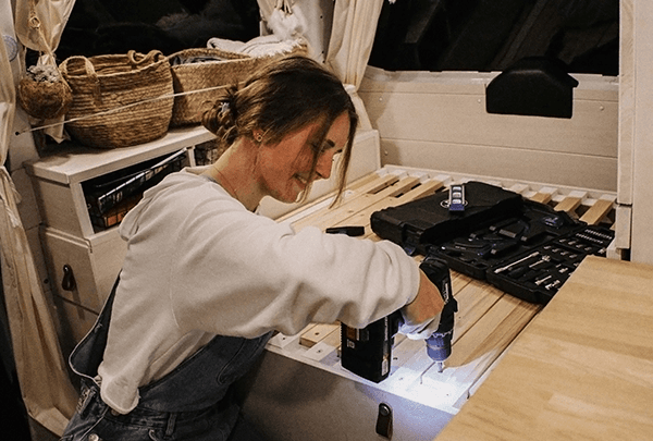 Jenni converting her van "Ellie" - @itsme.jenni 02/2022 A woman (@itsme.jenni) working inside her self-built camper van, using a power drill to assemble furniture. The van interior features cozy decor and organized tools, reflecting her journey of transforming a VW T5 into a mobile home.