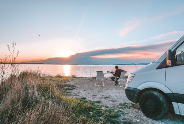 Ken playing guitar in front of their box truck - @offthemainroad_ 08/2021 Man sitting by a serene lakeside at sunset, using a guitar, next to a parked white van with the door open. The scene captures a peaceful evening with a vibrant sky reflected in the water, wild grass in the foreground, and birds flying overhead.