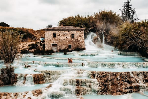 Exploring the thermal springs of Saturnia, Italy - @h.i.p.p.i.e_l.i.f.e 09/2022 Steffi and Pascal (@h.i.p.p.i.e_l.i.f.e) sit together in the cascading thermal pools of Saturnia, Italy, surrounded by milky turquoise water and lush vegetation. Behind them, a rustic stone building and a natural waterfall add to the picturesque scene. The layered terraces of steaming water flow gently down the hillside, capturing the peaceful, natural beauty of this iconic hot spring destination during their family vanlife journey.