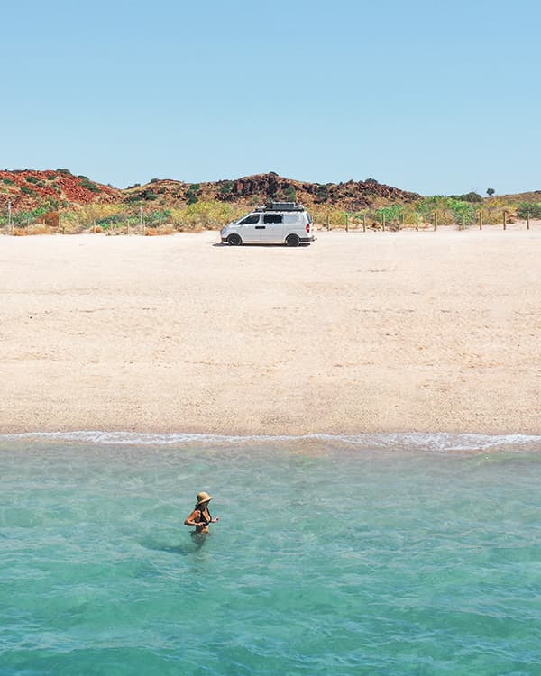 Hearsons Cove is real treat in the heat of Karratha, WA - @saltyvanlife 09/2022 Self-converted Hyundai van parked on a sandy beach at Hearsons Cove in Karratha, Western Australia, with Jade from @saltyvanlife wading in clear turquoise water against a backdrop of rugged red terrain.