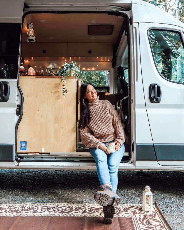 Emily in her RAM Promaster - @emsvanlife 08/2021 Emily Jay, a solo female traveler, sitting in the open doorway of her white RAM van, which is set up for vanlife. The van interior is decorated with string lights and plants, creating a cozy atmosphere. Emily is holding a colorful mug, wearing a brown sweater and jeans, and is parked in a wooded area, capturing the essence of her vanlife experience in Canada.