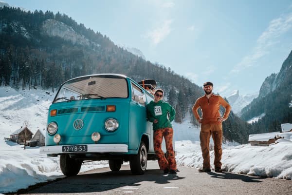Milene & Yuri facing cold temperatures with their VW T2 "Alexine" - @mygrations.nl 02/2022 Milene and Yuri standing beside their turquoise Volkswagen T2 van, Alexine, on a snowy mountain road. The backdrop features snow-covered mountains and a pine forest under a clear blue sky, capturing the essence of a winter adventure on their Silk Road journey. The image highlights van life, road trips, and exploration through remote, scenic landscapes in Europe.
