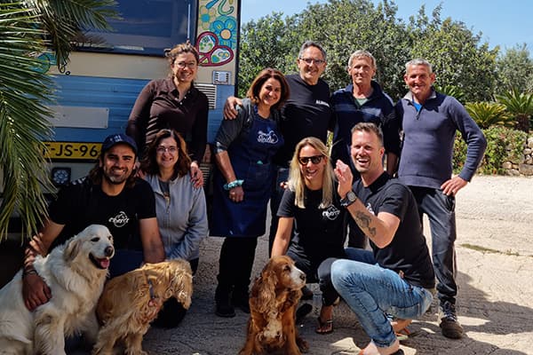 Visiting friends in Sicily - @travelcrazy.nl 09/2022 Renée and Joris from @travelcrazy.nl with their camper van Otto in Sicily, surrounded by their surrogate family and friends, smiling alongside several dogs in a sunny outdoor setting.