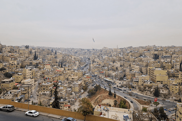 Panoramic view of Amman - paul.jaunegg 03/2023 the capital city of Jordan, showing densely packed beige buildings spread across rolling hills under an overcast sky. The large Jordanian flag rises above the skyline, symbolizing the city’s heritage and vibrancy. Photo by Paul Jaunegg.