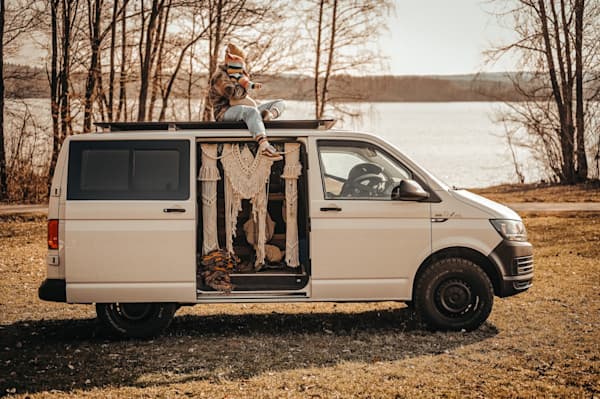 Steffi & her baby on top of their van - @h.i.p.p.i.e_l.i.f.e 09/2022 Steffi (@h.i.p.p.i.e_l.i.f.e) sits on the roof of a white VW T6 camper van with her baby in her lap, both warmly dressed in cozy layers. The van's sliding door is open, revealing a handmade macramé curtain hanging inside. Set against a peaceful lakeside backdrop with bare trees and soft golden light, the scene captures a quiet family moment in nature during their vanlife journey.