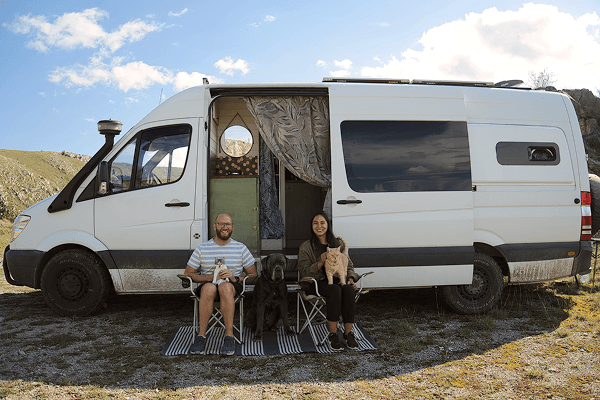 Our little family - @coyama.live 03/2023 A white Mercedes Sprinter camper van with its side door open is parked on a gravel clearing. In front of the van sit Barry and Tamara from @coyama.live, each on a camping chair with their pets. Barry holds Rocky the rescued kitten, their large black dog Leo sits between them, and Tamara holds their ginger cat Hamish. The van interior shows wood accents and patterned curtains, capturing a cozy vanlife scene during their long term travel journey.