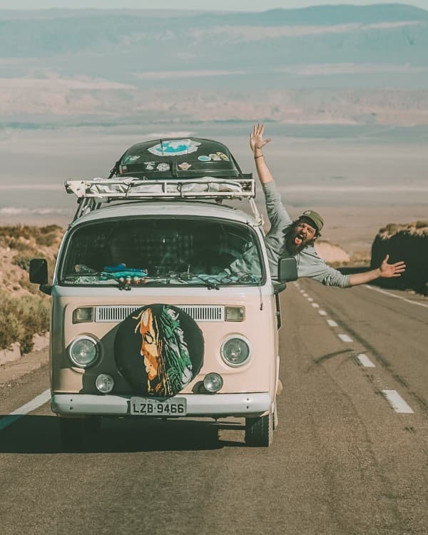 A great time on the road - @enjoytripbr 10/2020 A person joyfully extends their arms out of the passenger window of a vintage Volkswagen van traveling on a desert road. The van, adorned with stickers and a front spare tire cover featuring a wolf design, conveys a sense of adventure against a backdrop of distant mountains under a clear blue sky.