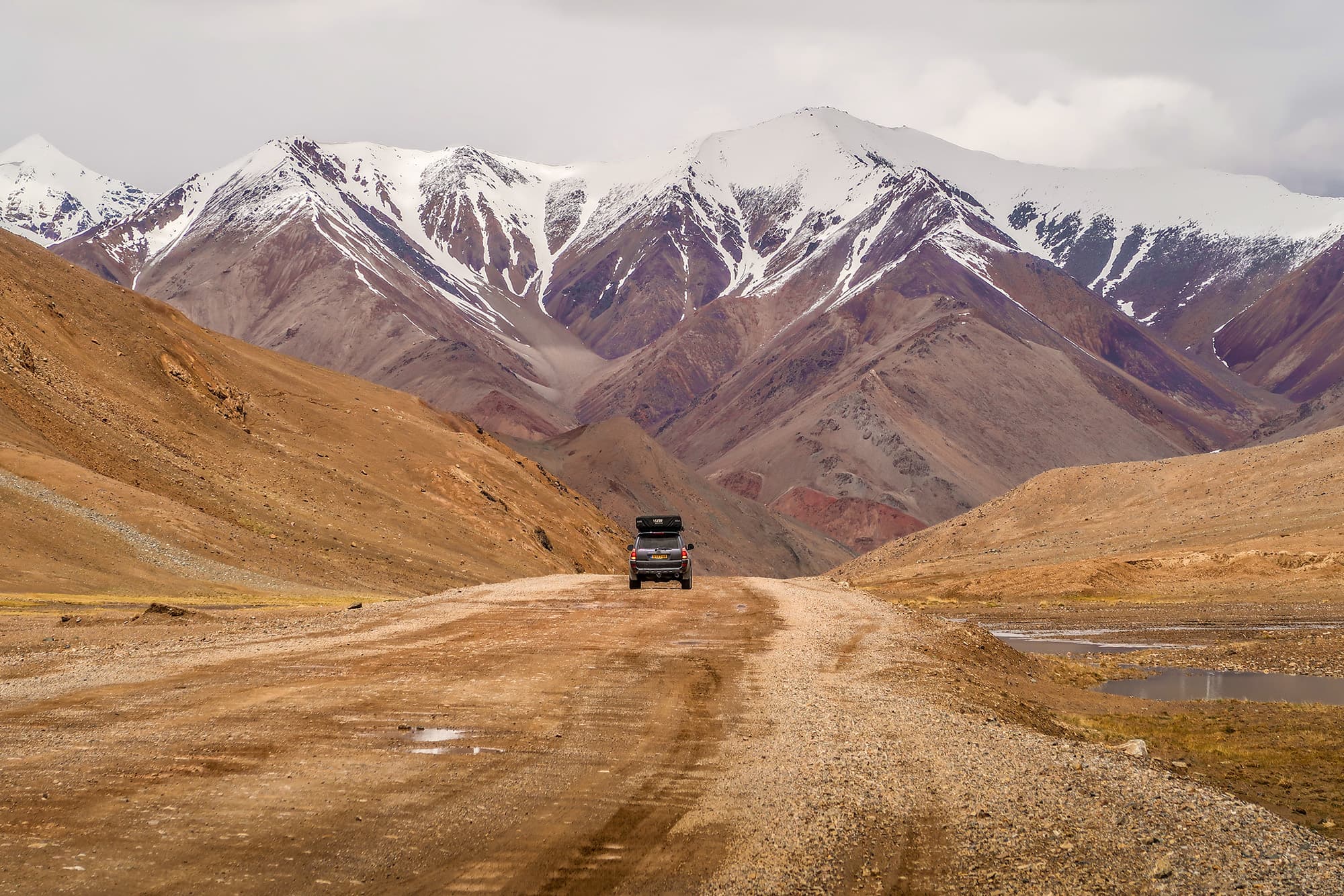 Driving the Pamir Highway in Tajikistan - @char.is.far 01/2025 A Toyota 4Runner with a rooftop tent, driven by Charelle and Marcel from @char.is.far, travels along the rugged Pamir Highway in Tajikistan. The vehicle is positioned on a dirt road surrounded by barren, rocky landscapes and dramatic snow-capped mountains in the background. This image captures the remote, high-altitude beauty of one of the world’s most iconic overland routes, part of their journey from Europe to Asia.