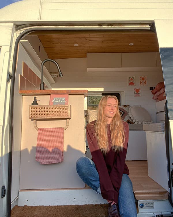Tamzin in the doorway of her converted Ford Transit van - @tamzin_in_transit 06/2022 @tamzin_in_transit sitting in the doorway of her converted 2011 Ford Transit van, showcasing a cozy van life setup with a wood-paneled ceiling, a kitchenette with pink tiles, a farmhouse sink, and stylish decor. The open door reveals a bright and inviting interior, reflecting a minimalist and adventurous lifestyle.