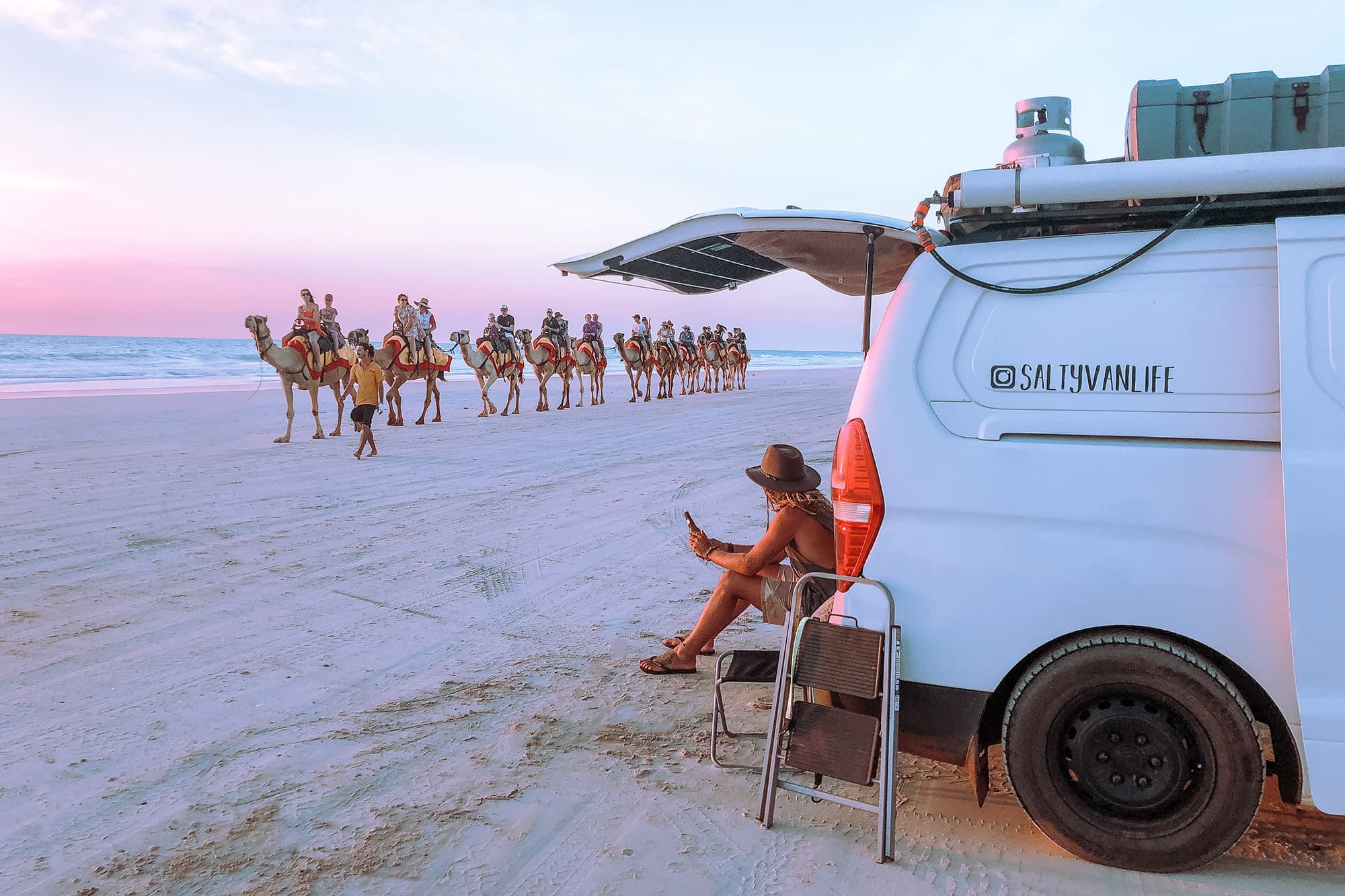 Enjoying the sunset at Cable Beach, WA - @saltyvanlife 09/2022 A man, Jake from @saltyvanlife, is sitting beside a self-converted van on Cable Beach at sunset, watching a caravan of camels walking along the sandy shoreline in Broome, Western Australia.