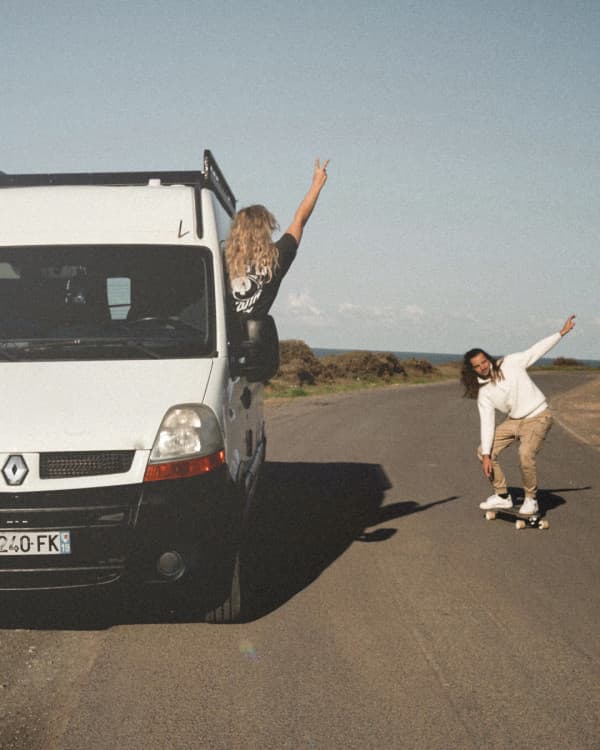 Making the open road a playground - @irisandmax 06/2022 A young couple, @irisandmax, enjoying vanlife with their converted Renault Master van. The woman leans out of the van, raising a peace sign, while the man skateboards alongside on a scenic road with clear skies and nature in the background. Vanlife adventure captured in motion.