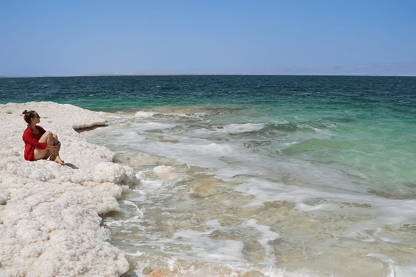We were lucky to find a wild beach at the Dead Sea - @kkdoo_pl 03/2023 Kasia from kkdoo_pl sits on a natural salt formation along the Dead Sea shore in Jordan, gazing out over the turquoise and emerald waters under a clear blue sky. The bright white salt crust contrasts with the vivid sea colors, capturing the beauty of vanlife travel, overlanding, and exploration in the Middle East.