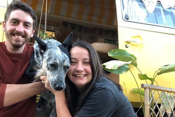 We might be on the road for a few years - @wanderingotooles 09/2022 Solomon and Justine of @wanderingotooles smiling alongside their black-and-white dog in front of their vintage yellow Volkswagen Kombi van. The van's open door reveals yellow-and-white striped curtains and a cozy interior. Green plants are visible around the van, adding to the outdoor, van life atmosphere of their Australian adventures.