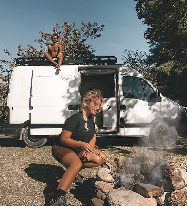 The fam! - @irisandmax 06/2022 A young couple, @irisandmax, enjoying vanlife at a campsite with their converted Renault Master van. The woman tends to a campfire, while the man sits on top of the van, surrounded by trees and nature, capturing the essence of outdoor living and vanlife adventures.