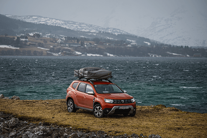 My Dacia Duster, which I converted into a camper - @hrvoje_juric 12/2022 Hrvoje Juric’s red Dacia Duster camper with a rooftop tent is parked beside a fjord in northern Norway during his winter vanlife expedition. Snow-dusted mountains and traditional red houses line the distant shoreline, illustrating the remote and scenic backdrop of his aurora borealis photography journey through Europe.