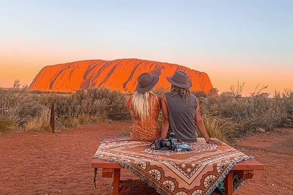 Watching the sunset at Uluru - @saltyvanlife 09/2022 Jade and Jake from @saltyvanlife sitting on a picnic table covered with a patterned blanket, watching the sunset over Uluru (Ayers Rock) in Australia’s red desert landscape.