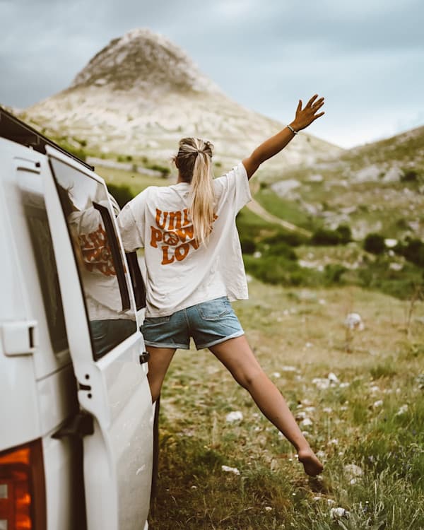 Embracing the Mountains - @h.i.p.p.i.e_l.i.f.e 09/2022 Steffi (@h.i.p.p.i.e_l.i.f.e) leans playfully out of the side door of a white camper van, wearing a loose shirt that reads “UNITY PEACE LOVE.” She stretches one leg toward the grassy ground while reaching her arm up, surrounded by rugged mountain scenery and a cloudy sky. A symbol of freedom and spontaneity on their family vanlife journey.