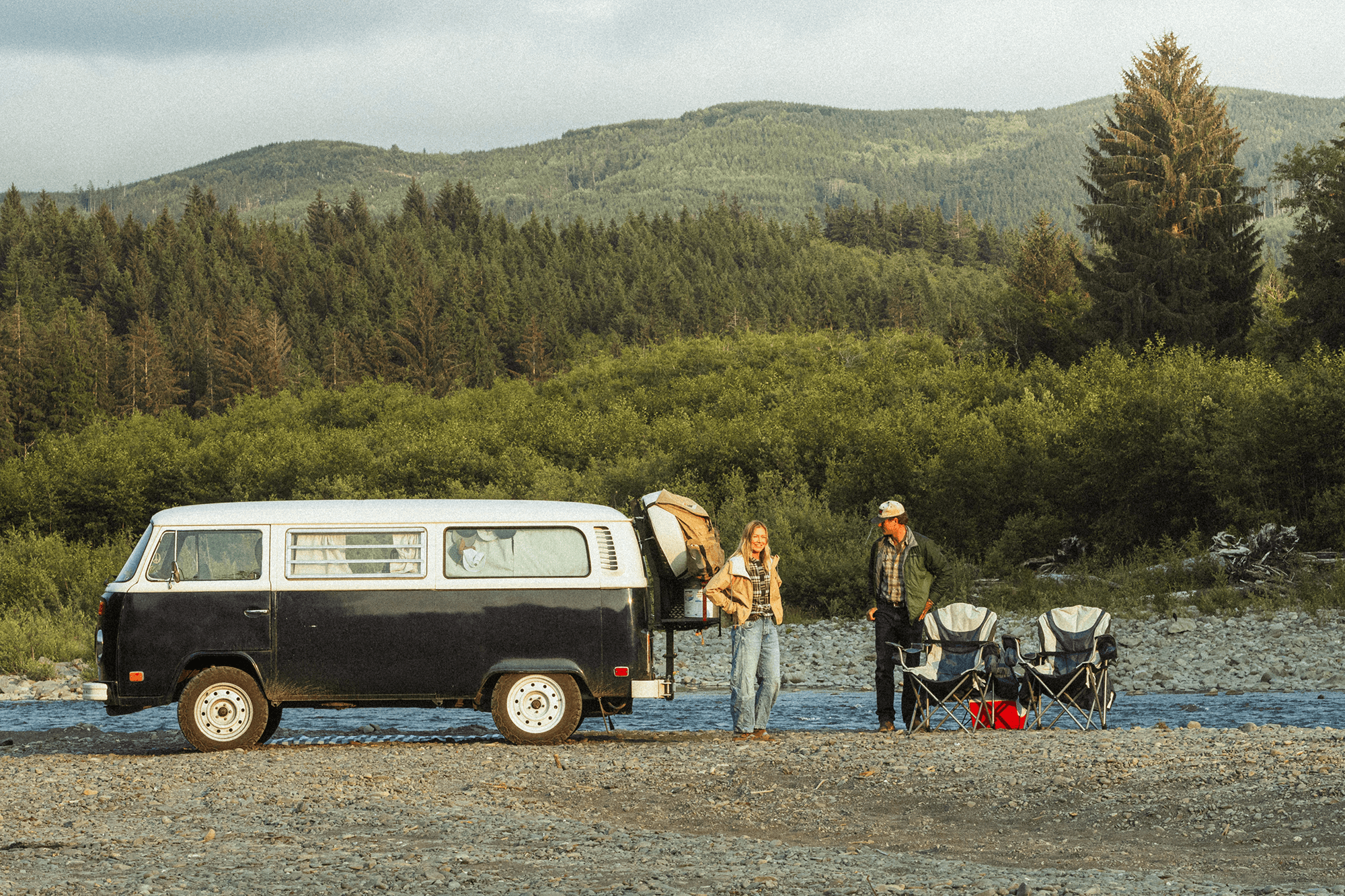 Teamwork is paramount - @wanderingnwonder 06/2025 Peter and Tara of @wanderingnwonder stand beside their black and white 1978 Volkswagen Bus named Wonder, parked along a peaceful riverbank surrounded by dense green forest and rolling hills. Two camping chairs and a red cooler are set up nearby, capturing the simplicity and serenity of their off-grid vanlife experience in nature during their cross-country USA journey.