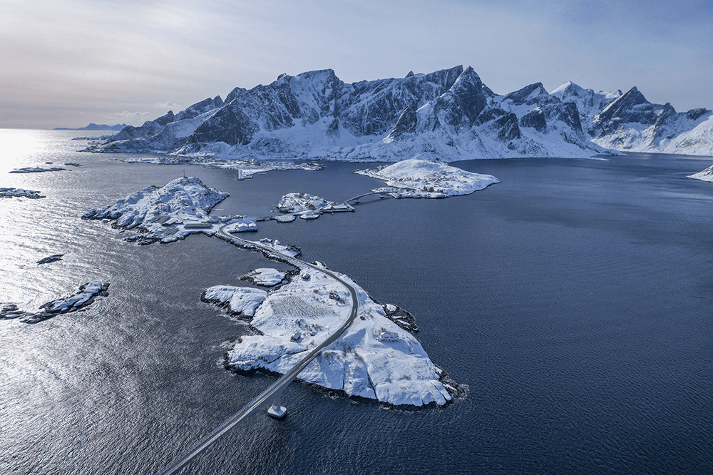 Winter on the Lofoten Islands - @hrvoje_juric 12/2022 Aerial view of the Lofoten Islands in Norway captured by Hrvoje Juric during his winter overland journey, showing a winding coastal road over snow-covered islands leading toward towering Arctic mountains, part of his Dacia Duster camper expedition to photograph the aurora borealis.