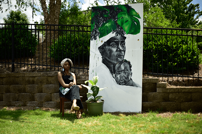 Allison & one of her artworks - driftwoodartwork 06/2025 Charcoal artist Allison Leah Thompson sits on a chair in a grassy yard next to one of her large-scale black and white artworks, which depicts a woman and child with green leaves above their heads. A potted plant is placed between her and the canvas. Known on Instagram as @idonthavegum, she creates and exhibits expressive portrait art while traveling the USA in her converted shuttle bus.
