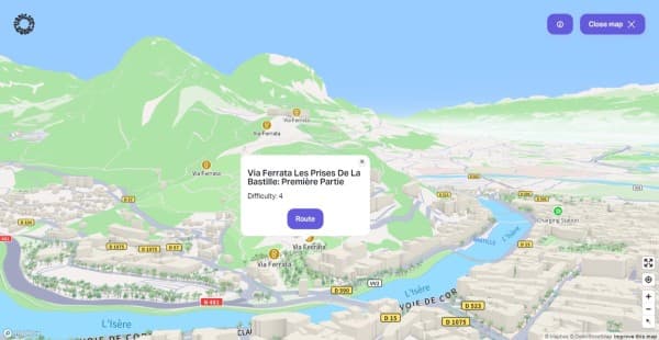 3D view of Grenoble, France with via ferratas - vanlifezone.com/map Interactive 3D map from Vanlifezone featuring the Via Ferrata Les Prises De La Bastille route in Grenoble, France. The map shows a detailed terrain of green mountains and the city's layout with buildings in relief, roads like D57 and D590, and the blue river L'Isère. A pop-up provides information on the Via Ferrata route, including the difficulty level, with a 'Route' button for navigation. Icons for Via Ferrata and a charging station are visible, aiding in adventure planning. The interface offers a 'Close map' feature and has Mapbox and OpenStreetMap attributions.