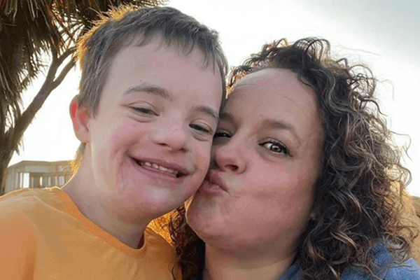 Helen & Leo - @cookingcampfireschromosones 05/2025 Helen and her son Leo from “Cooking, Campfires and Chromosomes” smile close together in a warm outdoor setting. Helen, with curly brown hair, kisses Leo on the cheek while he beams happily at the camera, wearing a bright orange shirt. Palm trees and soft sunlight frame the background, capturing a joyful, affectionate moment that reflects their loving bond, family travel life, and the spirit of inclusion and connection.