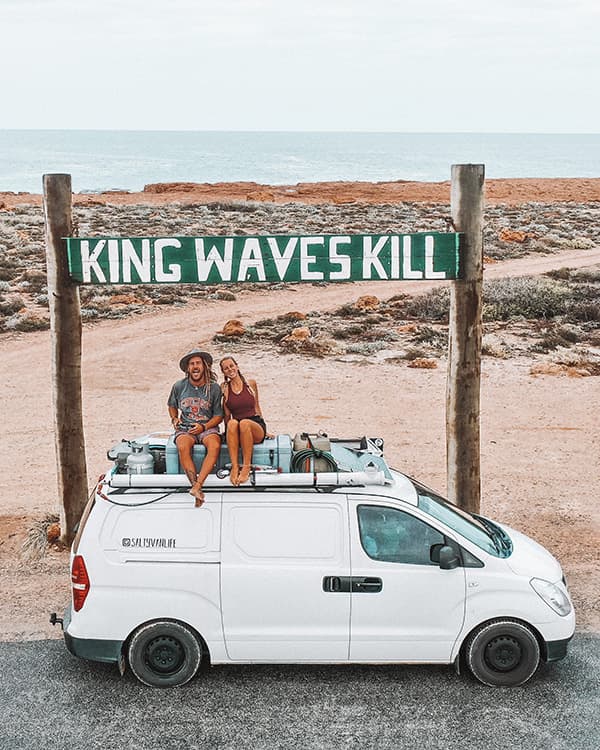 Jade & Jake under the "King Waves Kill" sign at Quobba Blowholes, WA - @saltyvanlife 09/2022 Jade and Jake from @saltyvanlife sitting on the roof of their self-converted van under the 'King Waves Kill' sign near rugged coastal cliffs at Quobba Blowholes, Western Australia.