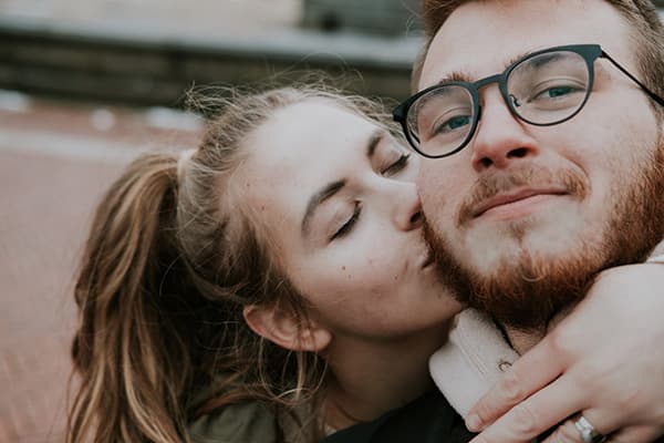 Madison & Brant - @harborhowls 09/2022 Madison, the author of this article and creator behind @harborhowls, leans in to kiss Brant on the cheek while wrapping her arm around him. Brant, with a beard and glasses, gazes softly at the camera. The background features a blurred outdoor setting, highlighting their warm and close connection.