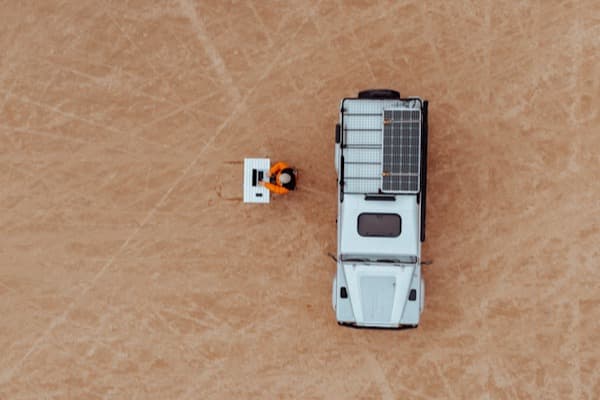 Space is very limited - @hypespeed 09/2022 @hypespeed's white Land Rover Defender, equipped with a solar panel and roof rack, is parked on an expansive, dry desert-like terrain. A person wearing an orange jacket and black beanie sits at a small white folding table, working on a laptop, creating a striking contrast against the vast, textured earth. The top-down drone perspective emphasizes the isolation and freedom of remote work, embodying the essence of van life and digital nomadism. The rugged surroundings and minimalist setup highlight the simplicity and self-sufficiency of an off-grid lifestyle.