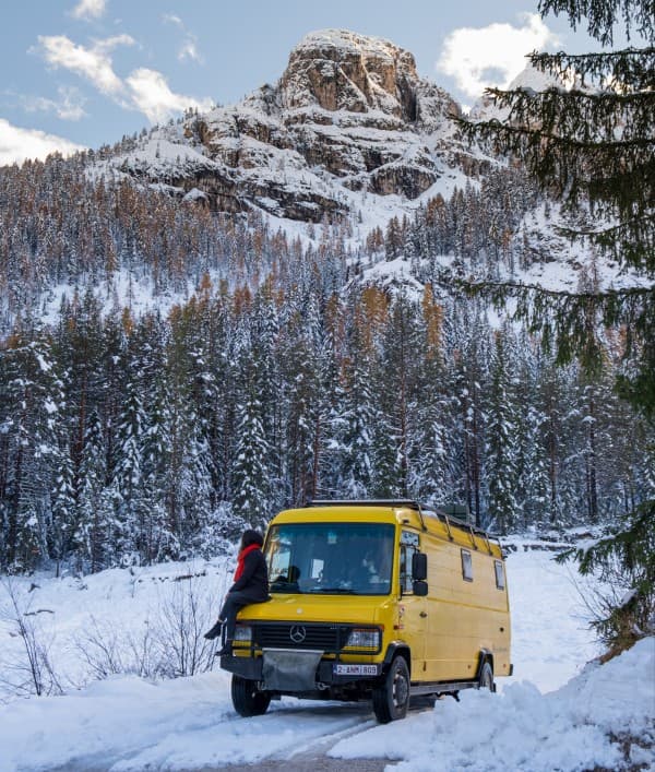 Vanlife in winter, a beautiful challenge - @our_orbit_vanlife 07/2024 Yellow Mercedes Vario 816D camper van from @our_orbit_vanlife parked in a snowy mountainous landscape with pine trees and a rocky peak in the background. One person, dressed in a red scarf and dark winter clothing, sits on the front bumper of the van, enjoying the serene winter scenery. The scene captures the essence of vanlife during winter, highlighting the adventure and beauty of nature.