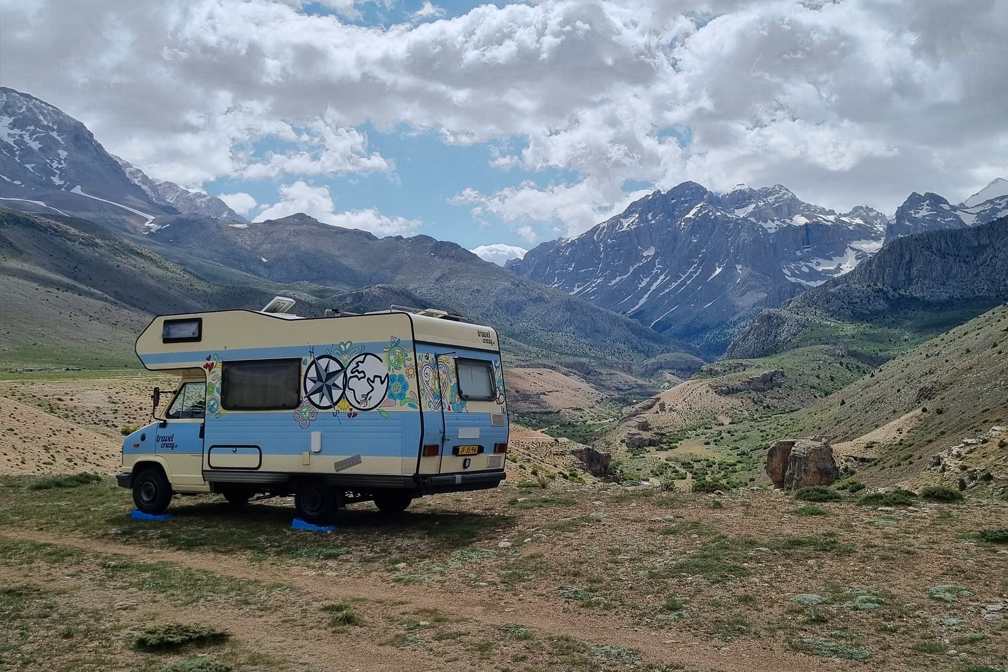 Otto parked in the rugged nature near Çamardı, Türkiye - @travelcrazy.nl 09/2022 @travelcrazy.nl's colorful camper van Otto parked on a grassy plateau near Çamardı, Turkey, with dramatic snow-capped mountains and a vast, rugged valley in the background under a partly cloudy sky, embodying the spirit of vanlife adventure in nature.