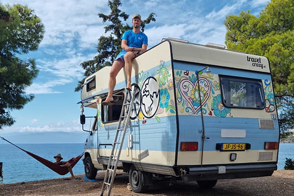 Enjoying the vanlife in Greece - @travelcrazy.nl 09/2022 @travelcrazy.nl’s camper van Otto parked near Ermionida, Greece, with Joris sitting on the roof accessed by a ladder and Renée relaxing in a hammock by the sea, surrounded by pine trees and a stunning coastal backdrop.