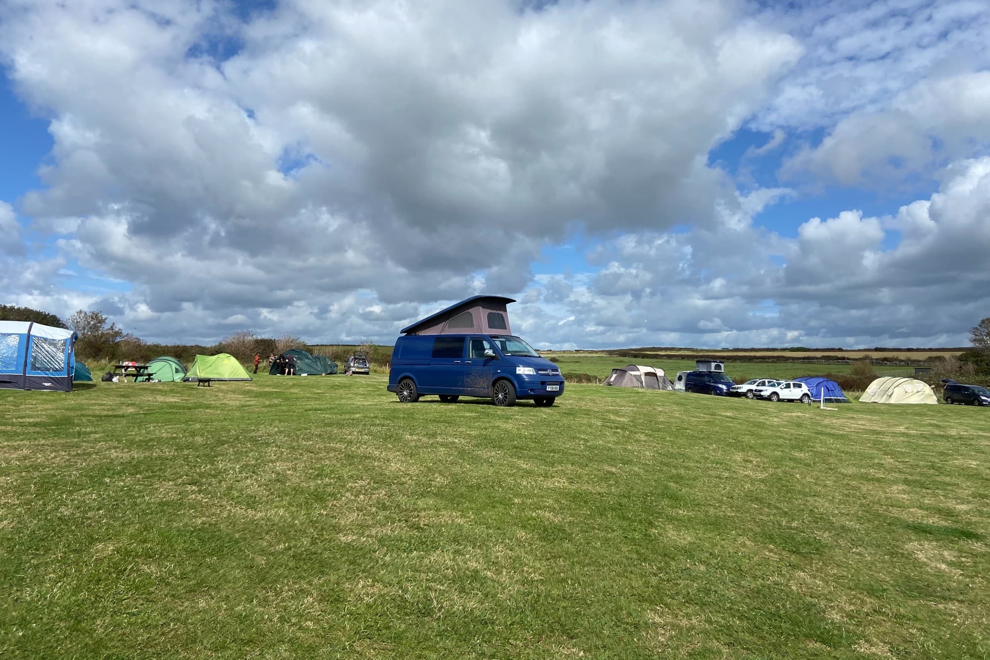 The Happy Camper, a Volkswagen T5 - @thehappycamper2020 10/2020 A blue Volkswagen T5 camper van with pop-up roof parked on a grassy field at a campsite in the UK, under a sky with dramatic clouds.