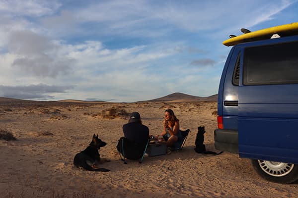 Jo, Georg with Momo & Nelly next to "Quest" - triptovantasia 06/2022 Jo and Georg from triptovantasia, with their dogs, sitting beside their blue VW T4 van in a desert landscape on the Canary Islands, enjoying a meal during their vanlife journey.
