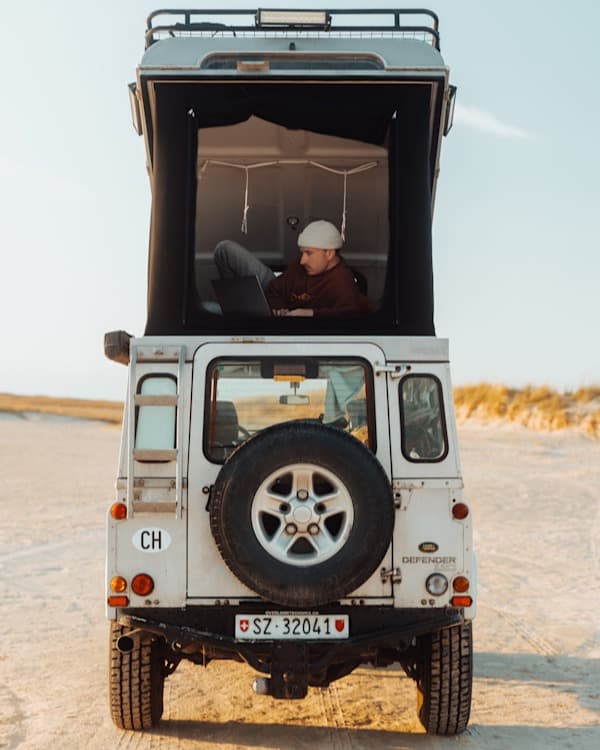 Roadoffice - @hypespeed 09/2022 @hypespeed's white Land Rover Defender with Swiss license plates (SZ 32041) is parked on an open sandy terrain under a clear sky. The vehicle is modified with a pop-up roof tent, which is fully extended. Inside the elevated sleeping area, a person wearing a beige beanie and a brown hoodie is reclining on their side, focused on a laptop. The scene captures the essence of van life and remote work, blending adventure and digital nomadism. The natural light and minimalistic background emphasize the rugged, off-grid lifestyle, making this a perfect representation of overlanding and mobile office living.