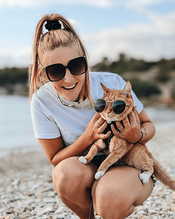 Hi, we're Chanti and Ollie - @chanti.and.ollie 12/2022 Chantal from @chanti.and.ollie kneels on a rocky beach smiling while holding her ginger cat Ollie, both wearing sunglasses, during a European vanlife adventure with their Opel Movano campervan.