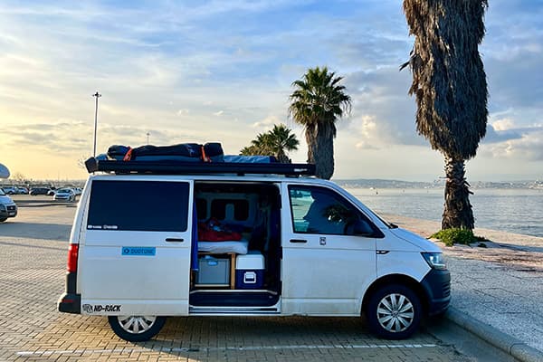 A commitment to a way of life - @van4life__ 04/2025 A white W T6 camper van belonging to Chris from @van4life__ is parked along a coastal promenade, facing the calm waters of a bay under a golden evening sky. The van’s sliding door is open, revealing a neatly packed interior with a cooler, storage boxes, and bedding. A surfboard bag is secured on the roof. Tall palm trees line the walkway, and the soft light reflects off the ocean, evoking the peaceful, minimalist lifestyle of vanlife on the Iberian coast.
