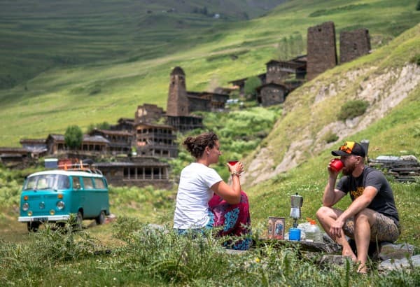 Milene & Yuri in Dartlo, Georgia with their van - @mygrations.nl 02/2022 Milene and Yuri enjoy a coffee break outdoors in a scenic mountain setting with their turquoise Volkswagen T2 van, Alexine, parked nearby. The background features the historic village Dartlo in Georgia with stone watchtowers and traditional buildings set among rolling green hills, reflecting a peaceful moment of van life and cultural exploration on their Silk Road journey.