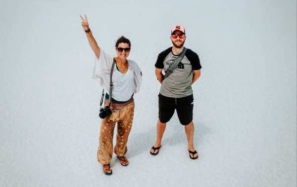 Milene & Yuri - @mygrations.nl 02/2022 Milene and Yuri from @mygrations.nl standing on a vast salt flat, with Milene making a peace sign and holding a camera, and Yuri standing beside her, both wearing casual travel clothing. The white, expansive landscape creates a minimalist backdrop, highlighting their adventurous spirit as they explore unique destinations on their journey along the Silk Road. The image captures a moment of travel, adventure, and cultural exploration.