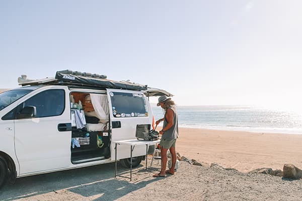 We do everything outside and its just how we like it - @saltyvanlife 09/2022 Jake from @saltyvanlife cooking on a portable stove next to their self-converted van parked by the beach, with an open ocean view and bright sunlight in Western Australia.