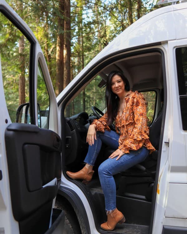 Emily in her Van - @emsvanlife 08/2021 Emily Jay from @emsvanlife sitting in the driver's seat of a RAM van in a forest setting, representing her vanlife journey. She is wearing a patterned orange blouse and jeans, with trees visible in the background, illustrating her outdoor lifestyle.