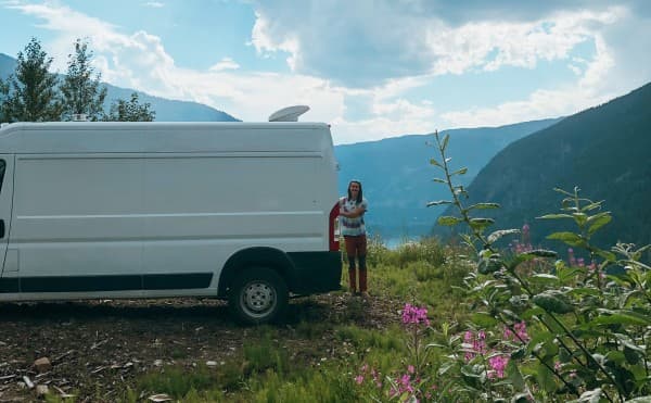 Alejandro next to their van - @georgiabroderick 08/2021 A person stands next to a white van parked amidst a wildflower-strewn landscape with a mountainous backdrop under a dynamic sky. The van's roof features a vent, illustrating the allure of mobile living and exploration in a serene, natural setting, perfect for outdoor and travel enthusiasts.