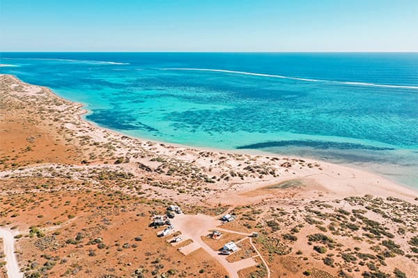 You are able to walk from the beach straight onto a coral reef - @saltyvanlife 09/2022 Aerial view of a camping area near the turquoise waters and coral reef of the Ningaloo Coast, Western Australia, surrounded by rugged coastal landscapes.