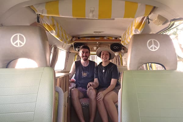 Solomon & Justine in their kombi - @wanderingotooles 09/2022 A young couple, Solomon and Justine of @wanderingotooles, sitting inside their vintage Volkswagen Kombi van. The van interior features light green seats with peace symbols, yellow-and-white striped curtains, and a cozy wooden setup in the background. The couple smiles warmly, showcasing their van life adventures in Australia.