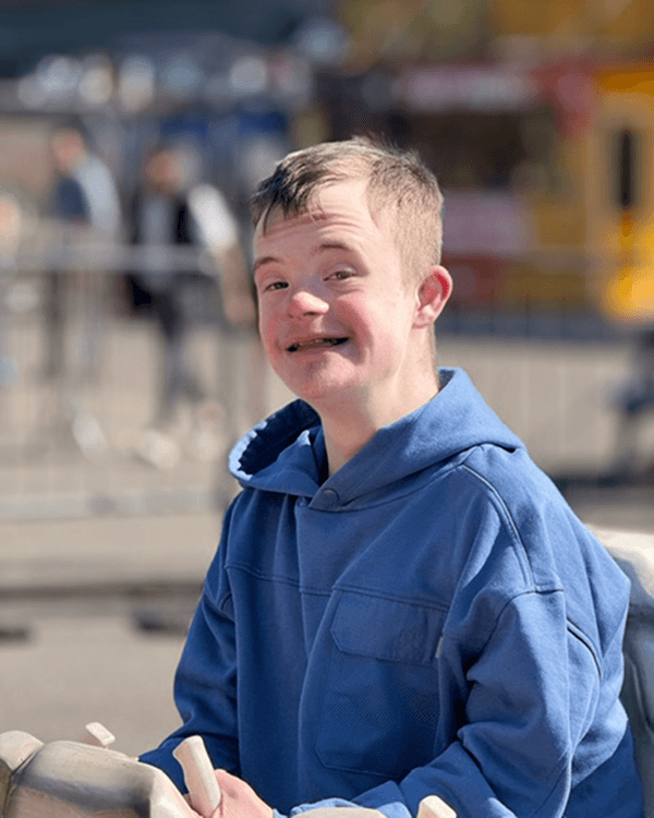 Leo spreads joy like sparks from a campfire - @cookingcampfireschromosones 05/2025 Leo from “Cooking, Campfires and Chromosomes” smiles warmly while sitting outdoors in a bright setting, wearing a soft blue hoodie. His hands rest on a sculpted ride or play structure, and the blurred background shows a sunny day with people walking and colorful stalls behind him. The image captures Leo’s joyful spirit and presence, reflecting themes of accessibility, inclusion, and daily life with Down syndrome.